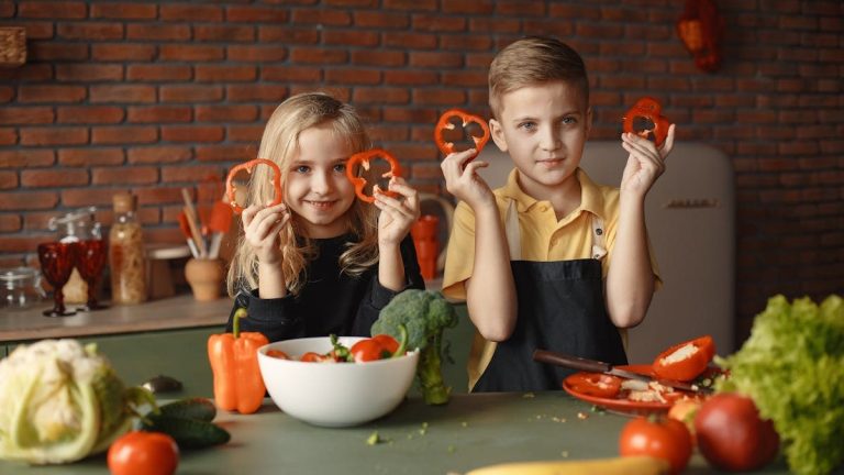 Two children holding capsicum slices while preparing vegetables in a cozy kitchen setting.